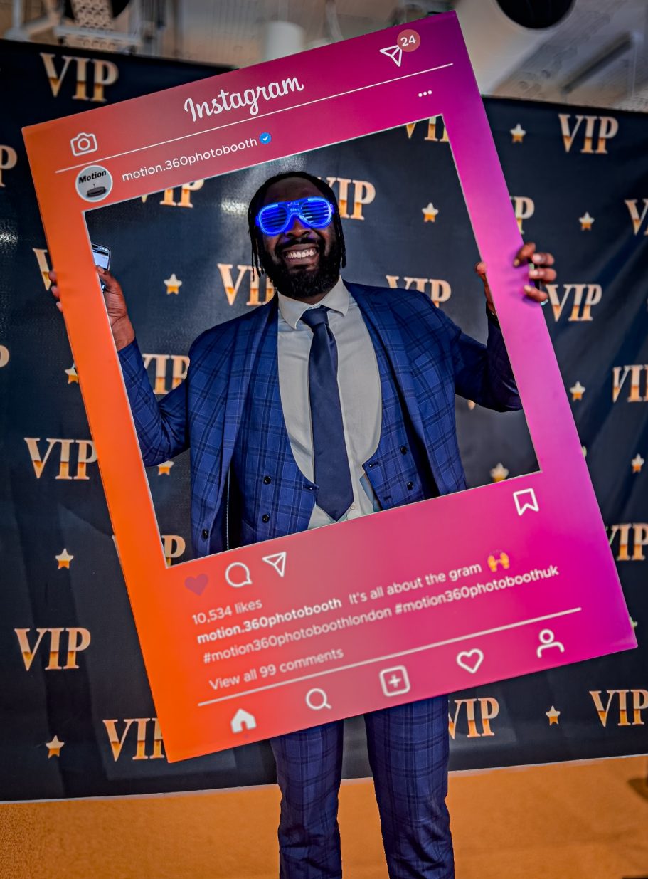 Man in a suit holding a large Instagram frame, wearing sunglasses and smiling.